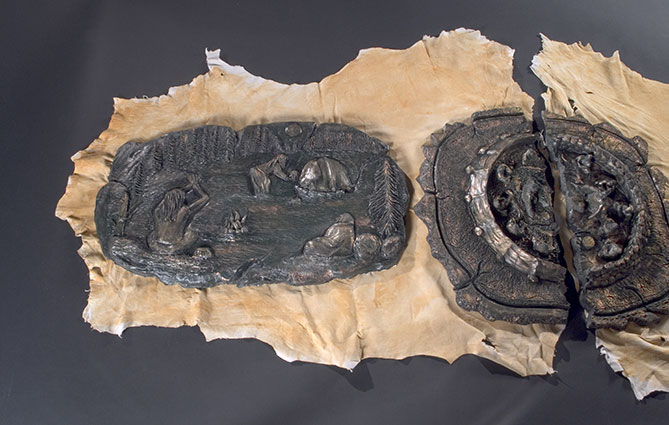 The left side of the work shows some aspects of traditional Innu ceremonies, such as a sweatlodge. Here the figures depicted are in a contemplative mood. By a fire, a shaman interprets the signs the fire has left on a caribou shoulder blade. The patinated plaster provides a bronze effect: greenish tones embellished with golden highlights. The set of sculpted pieces lies on two dyed skins with roughly ripped edges. The way these two elements play off each other gives rise to a palpable tension between two separate universes which each have their own codes and features. The contrast between these two worlds is heightened by the attitude of the figures depicted: the enraged priest and the contemplative shaman.
